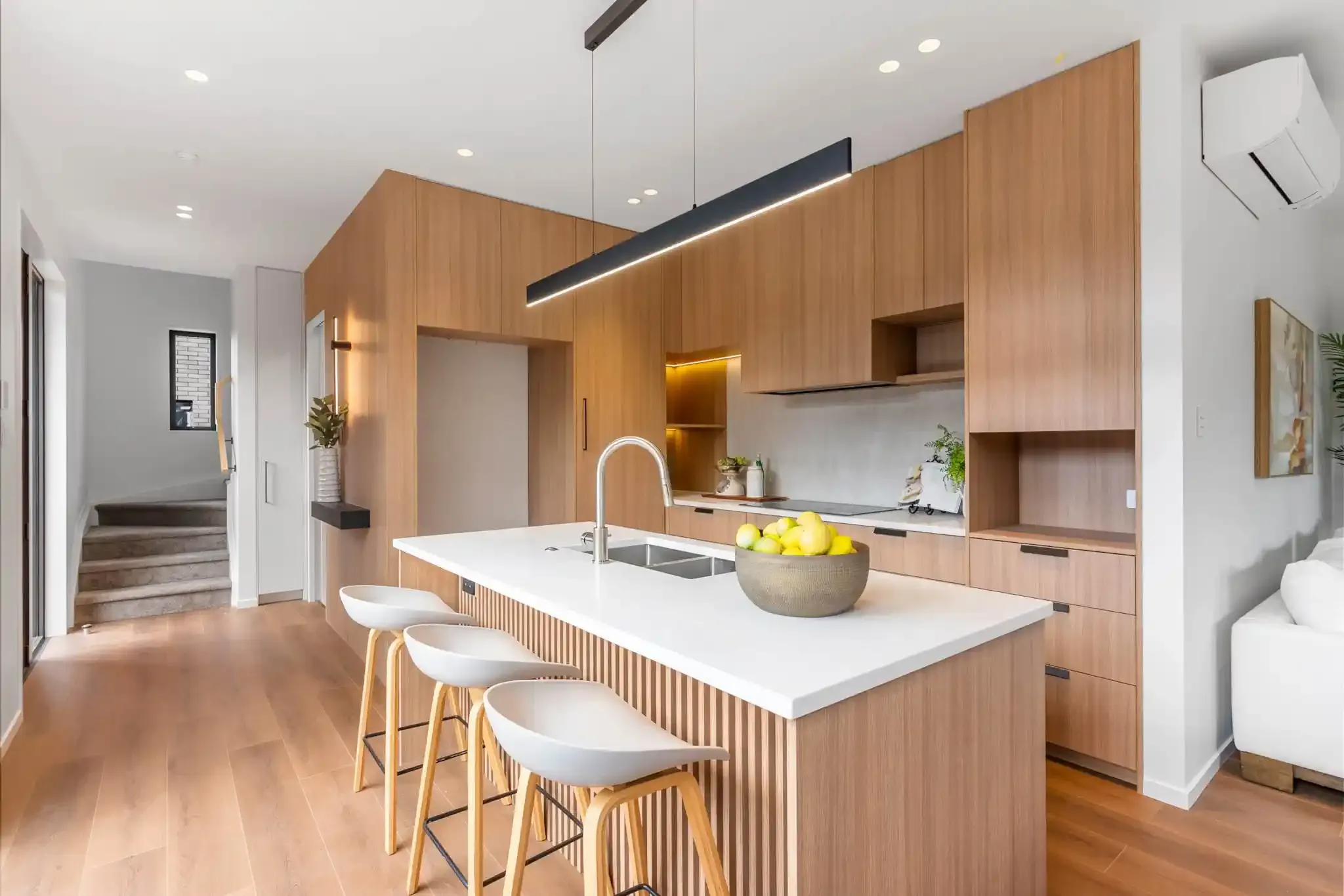 A kitchen with oak cabinetry and a white stone island on a fluted base, with the staircase and entry visible beyond.